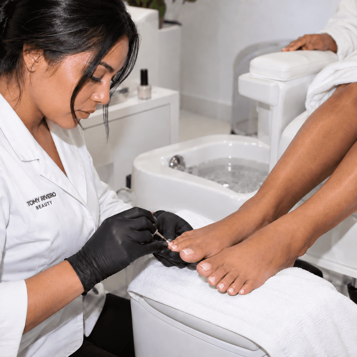 Pedicure specialist’s black-gloved hands applying polish to a client’s toenails in a bright treatment room